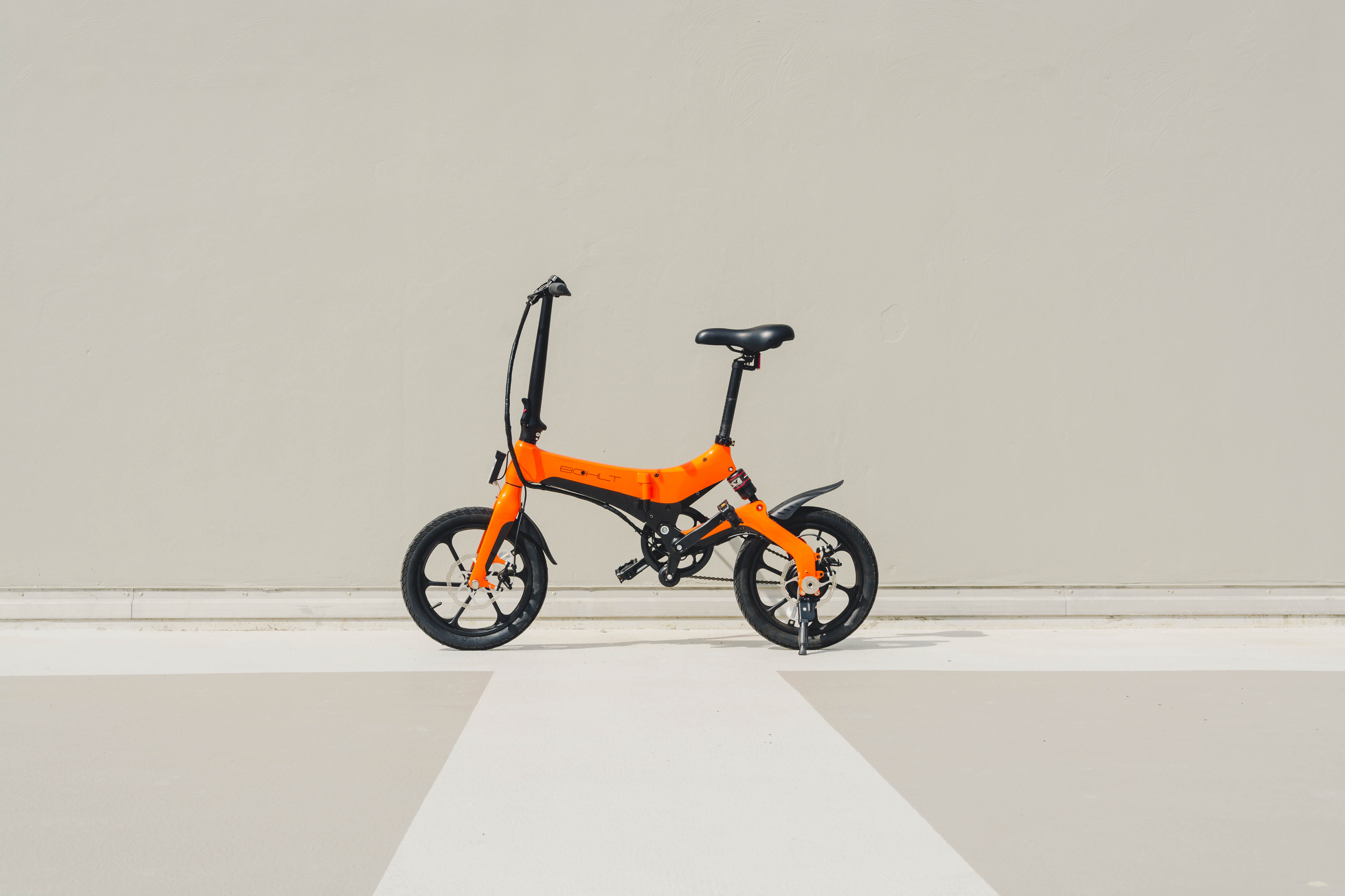 An orange folding bicycle is parked on a light-colored floor against a light gray wall.