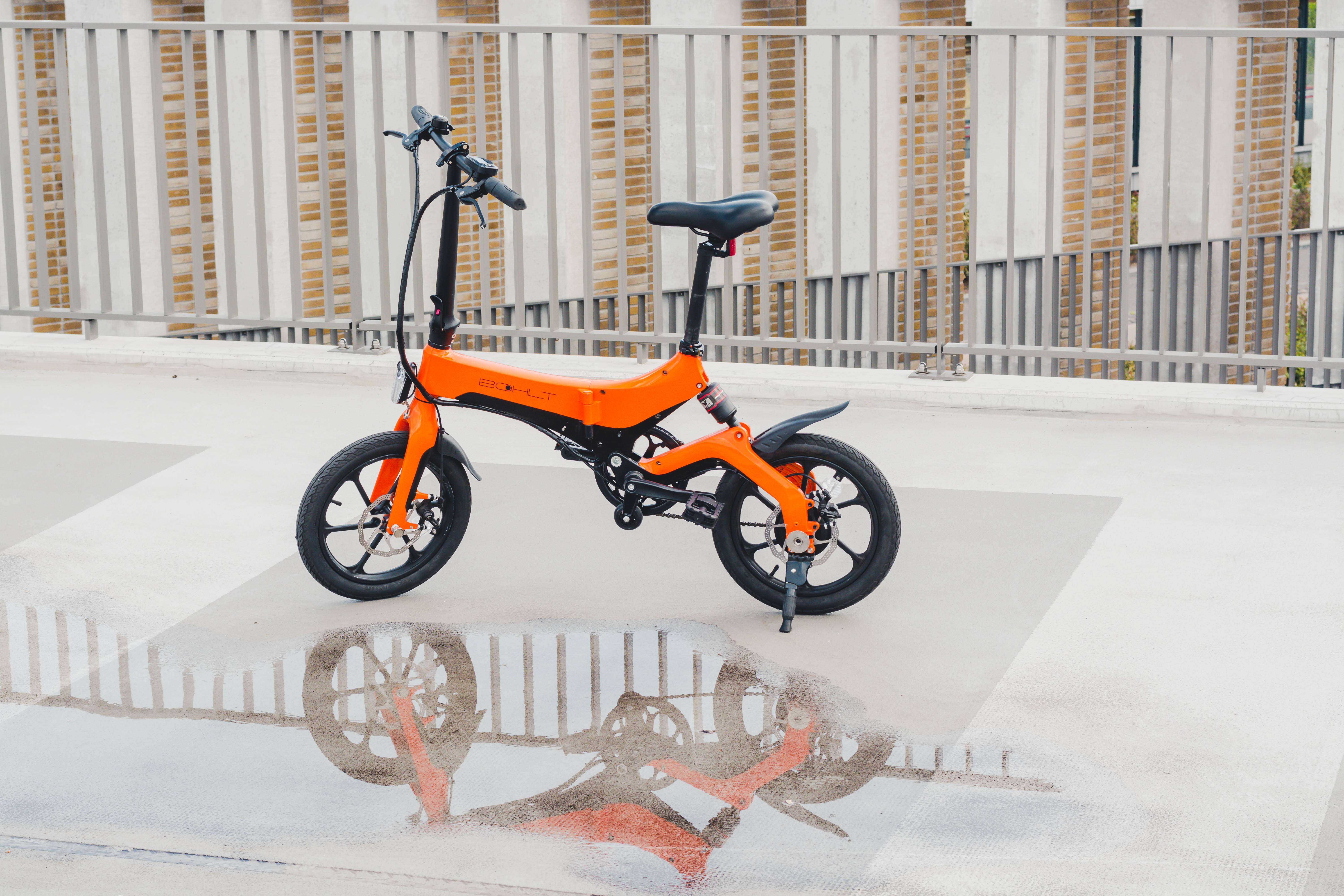 An orange folding electric bike stands on a wet surface with reflections.