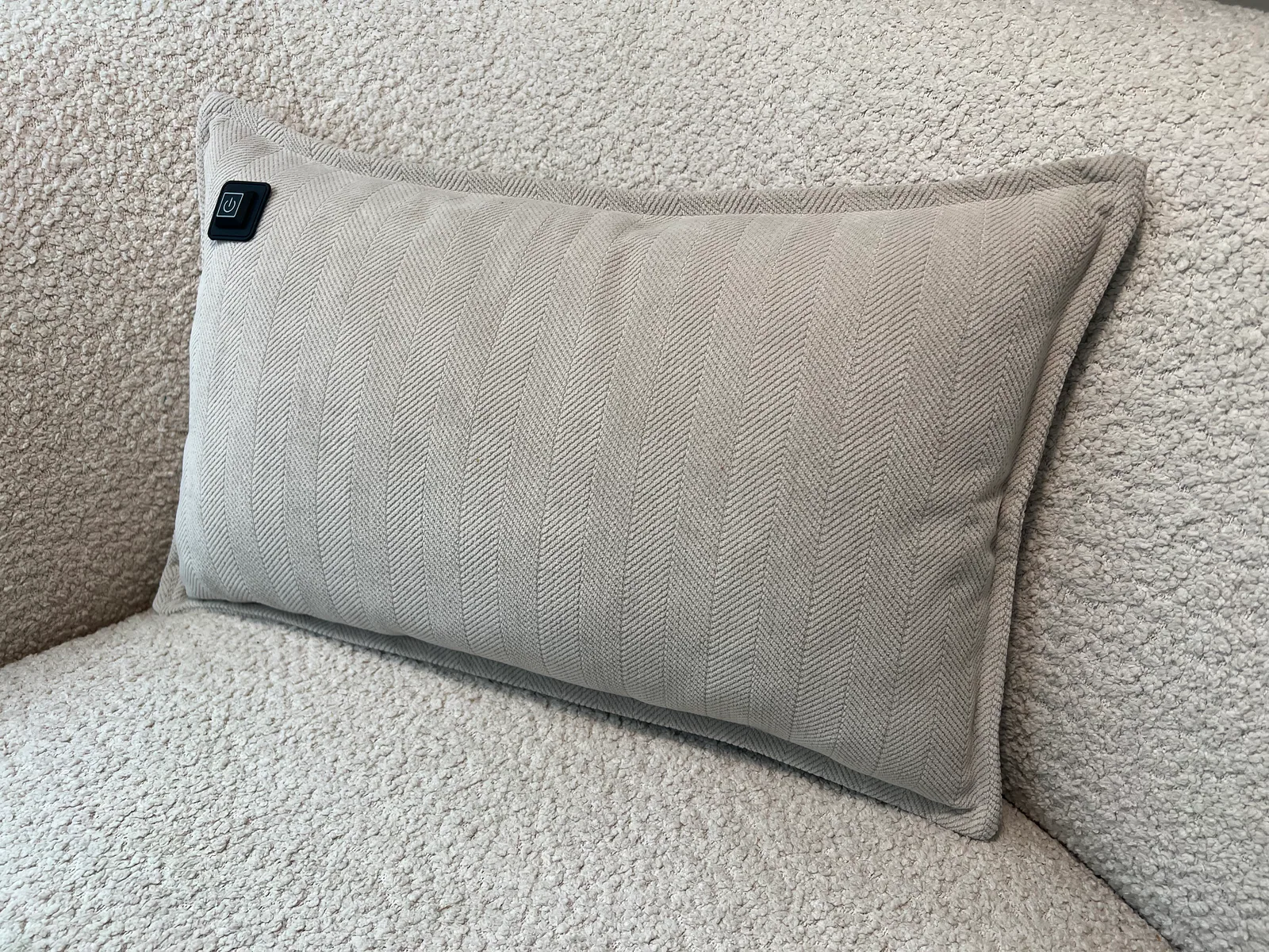 Ein hellgraues Kissen mit Fischgrätmuster liegt auf einem cremefarbenen Bouclé-Sofa. Rechts oben ist ein schwarzes Power-Symbol.