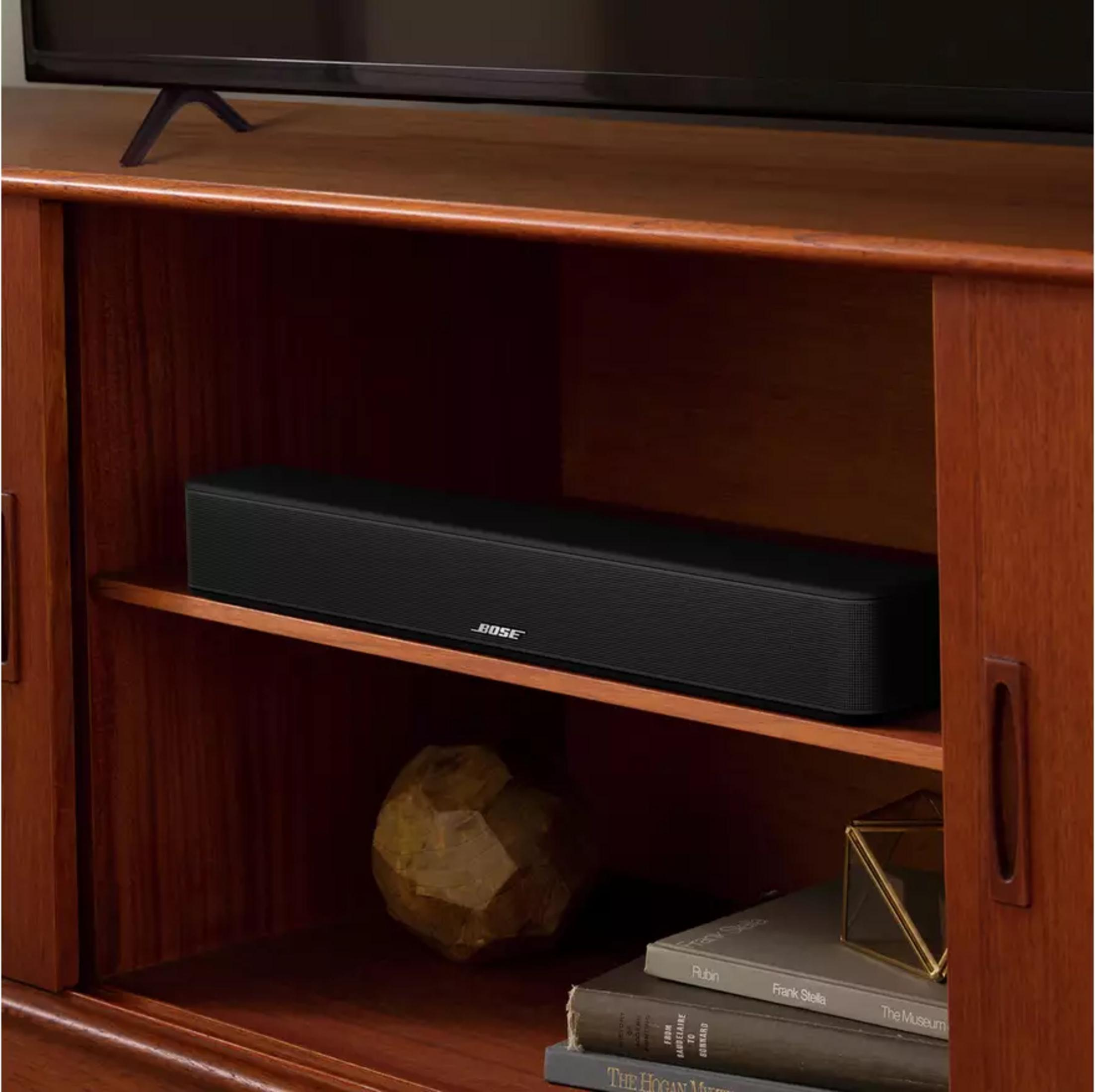 Barra de sonido Bose negra en estante de madera con libros y decoración.
