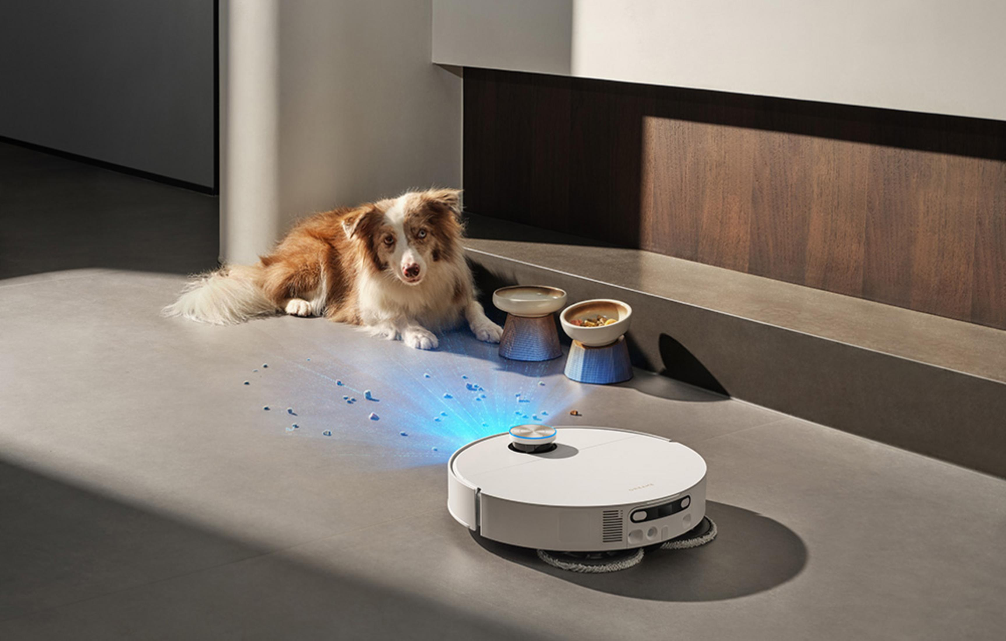Un cane giace vicino a un robot aspirapolvere che emette luce blu e sparge detriti.