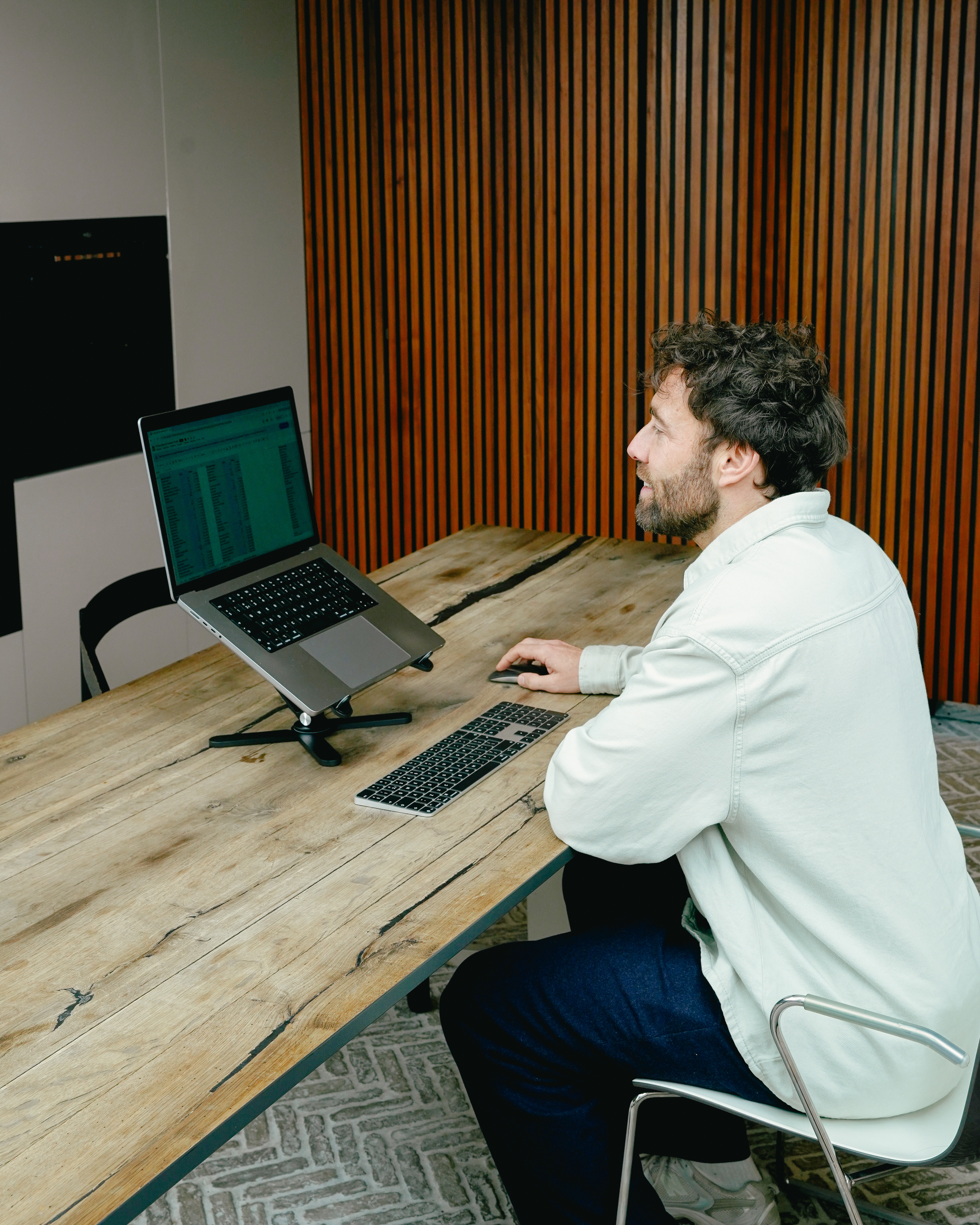 Homme barbu assis à un bureau en bois, travaillant sur un ordinateur portable avec clavier et souris.