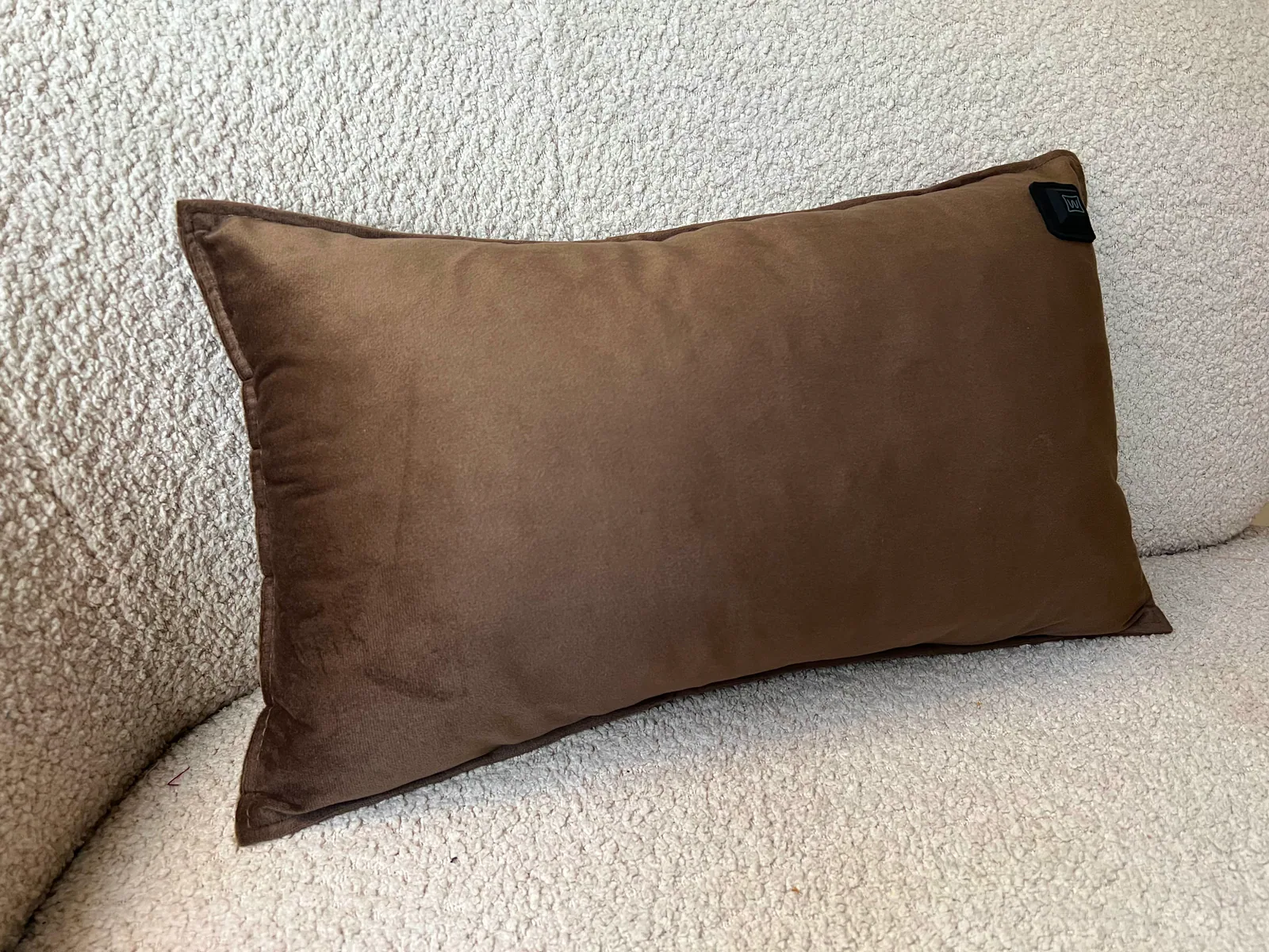 Ein braunes Samtkissen liegt auf einem cremefarbenen, flauschigen Sofa. Oben rechts ist ein kleines schwarzes Etikett.