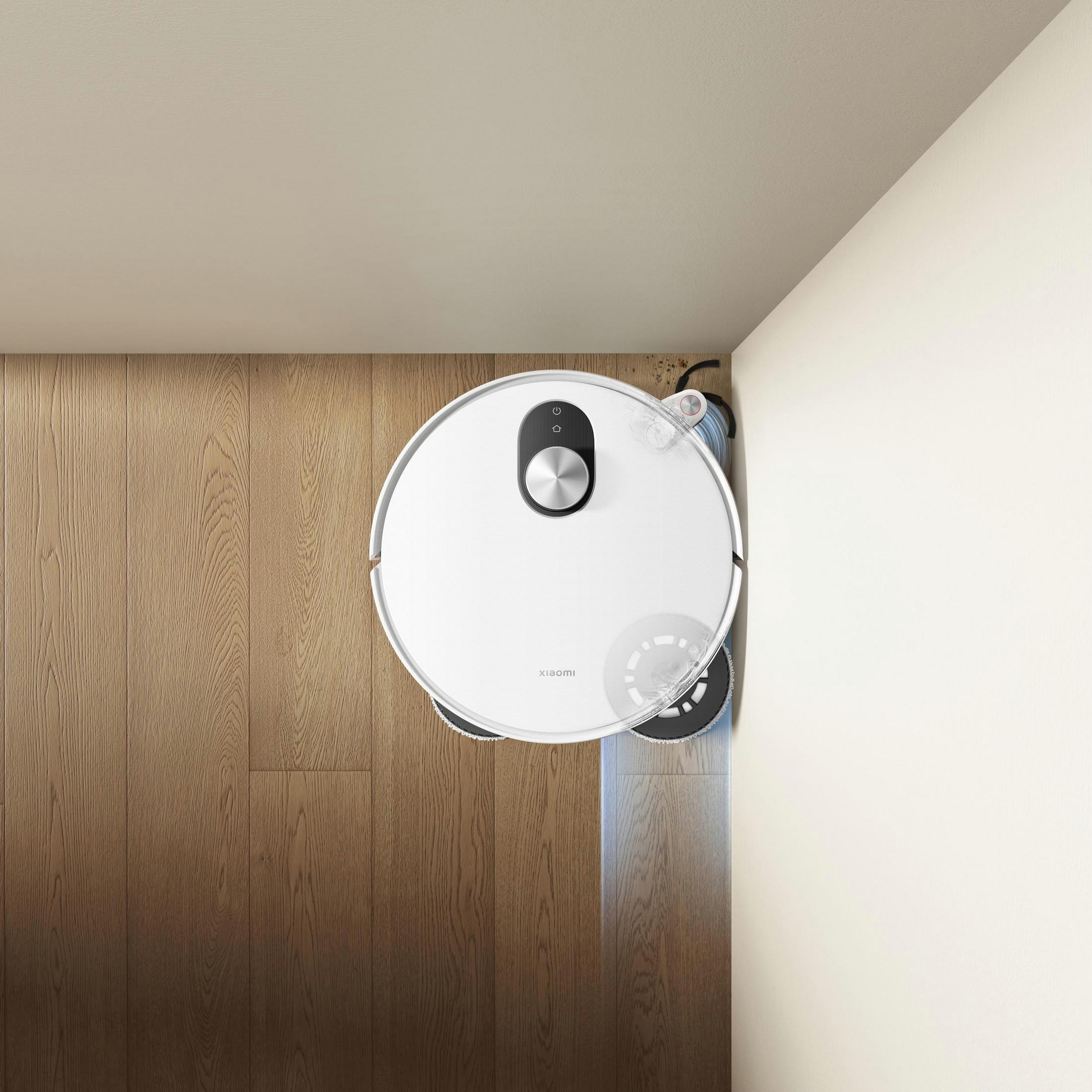 A white robot vacuum cleans a wooden floor next to a white wall.
