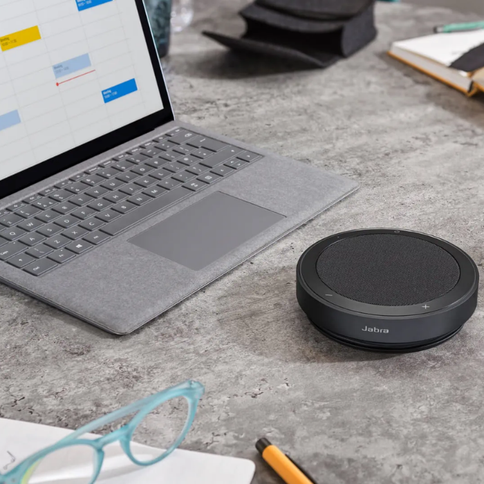 Ein Laptop, ein Jabra-Lautsprecher, eine Brille und ein Stift liegen auf einem Schreibtisch.