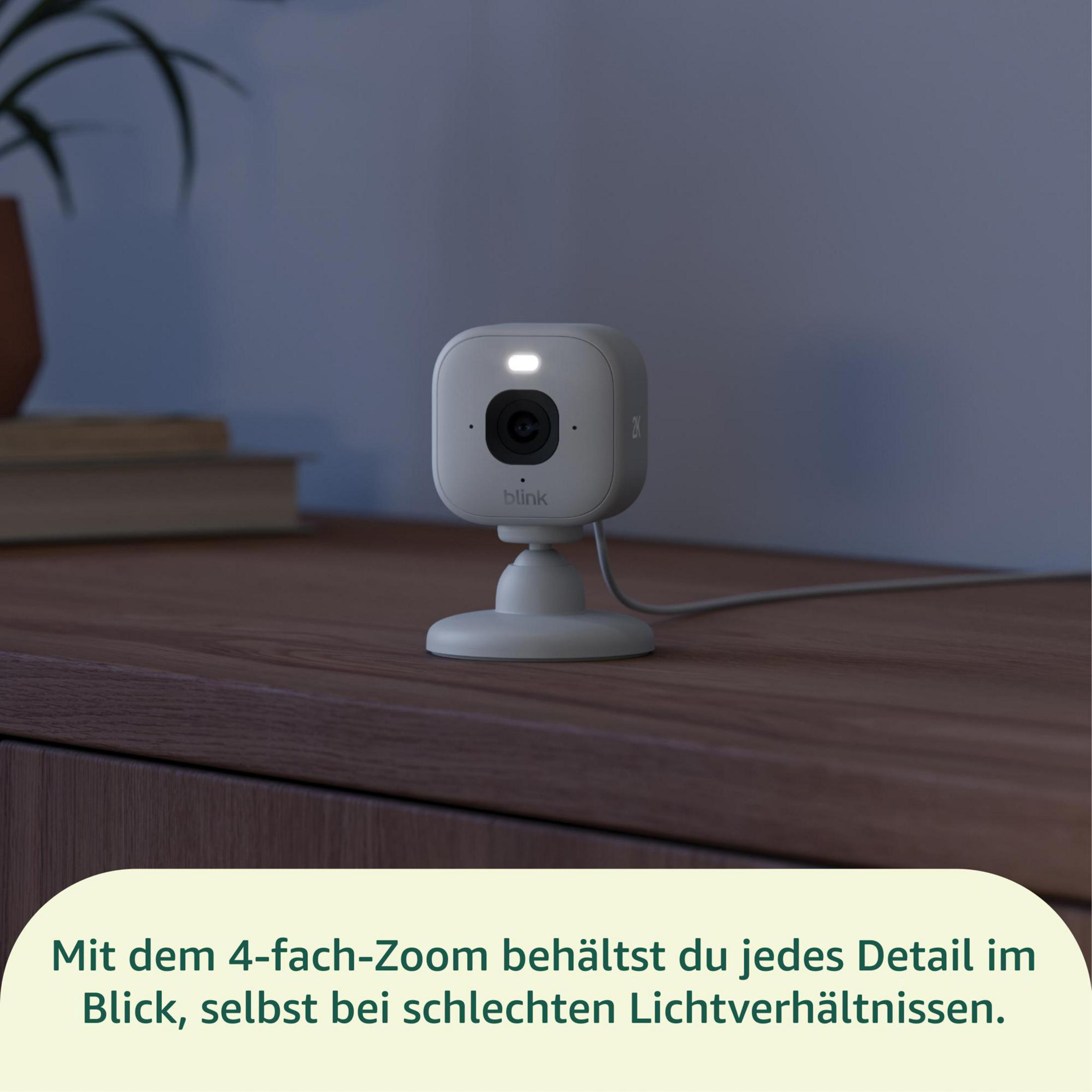 Eine weiße Blink-Kamera mit einem Licht steht auf einem Holztisch. Links sind Bücher.
