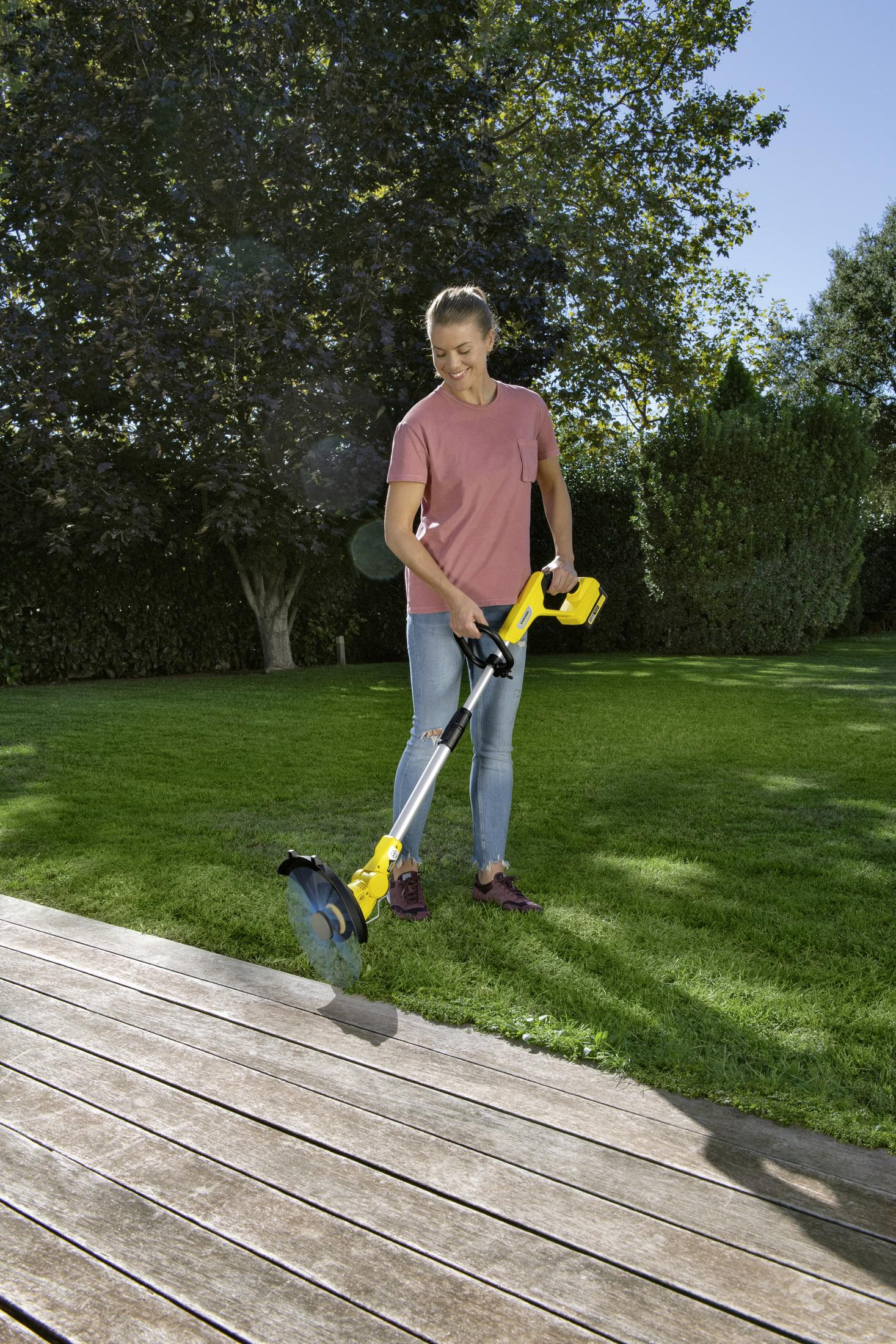 Frau im rosafarbenen T-Shirt trimmt Rasen mit gelbem Gerät auf Holzdeck mit Garten im Hintergrund.