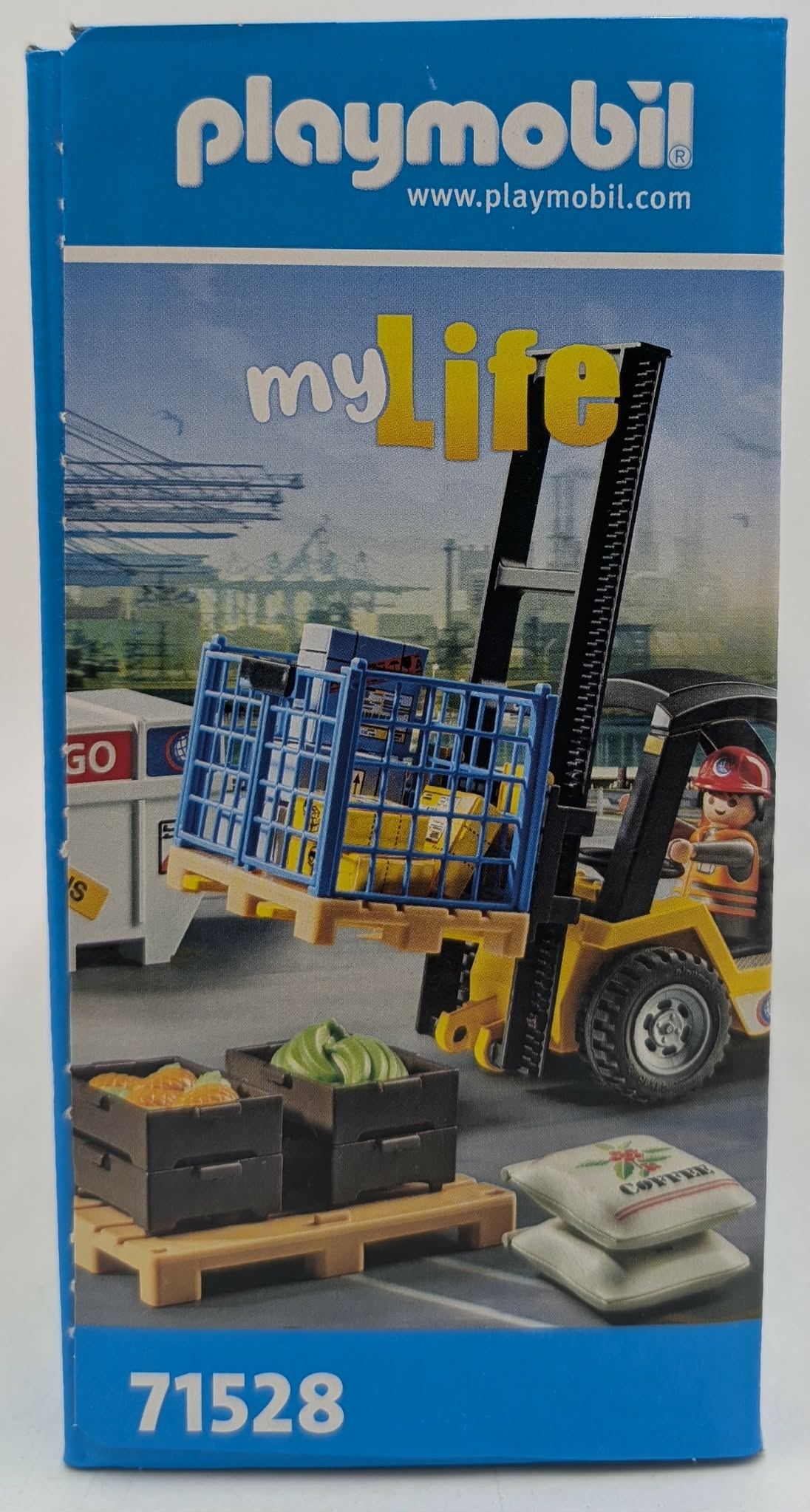 Carrello elevatore Playmobil solleva cassa blu con scatole gialle. Vicino ci sono casse di cibo e sacchi di caffè.