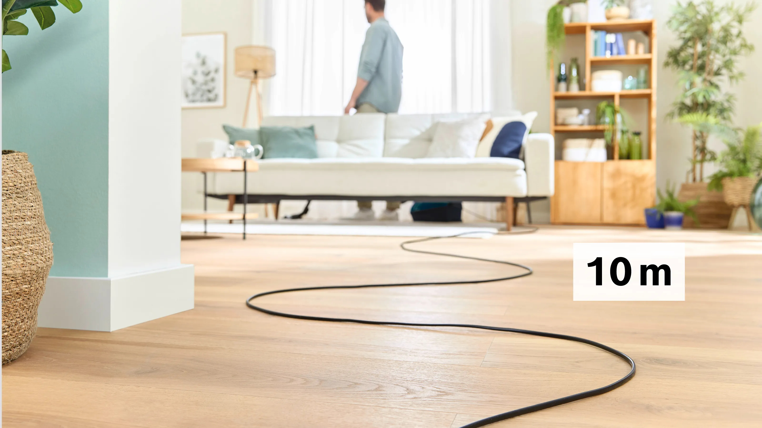Ein schwarzes Kabel liegt auf einem Holzboden. Ein Mann steht vor einem Sofa.