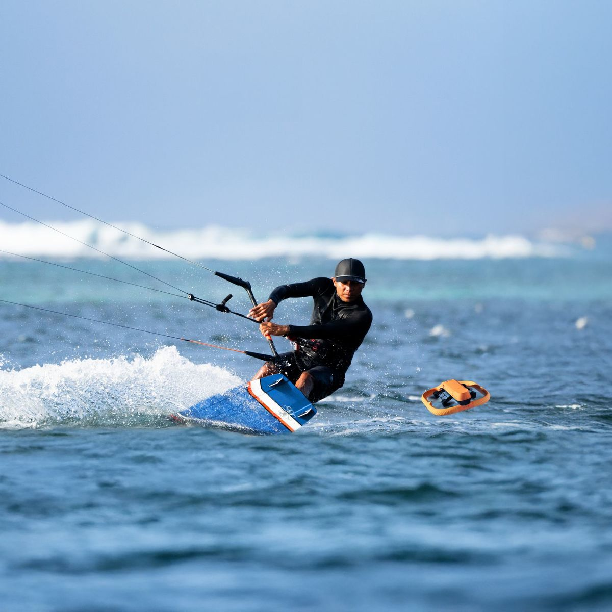 Kiteboarder w czarnym piance na niebieskiej desce surfuje po błękitnej wodzie, w pobliżu pomarańczowy przedmiot.