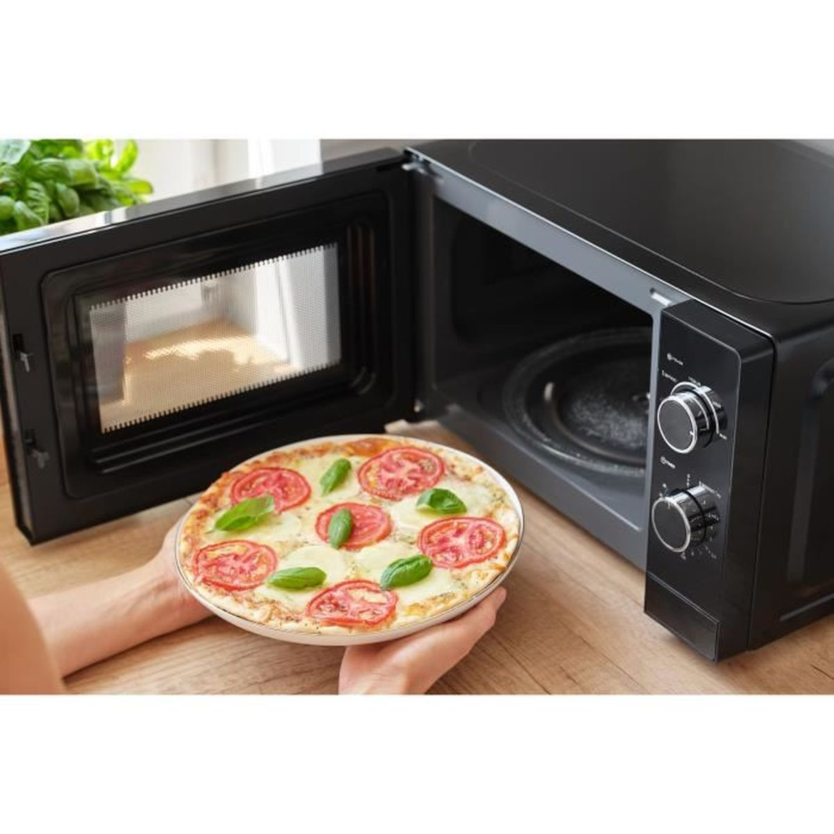 Una persona tiene una pizza Margherita davanti a un forno a microonde nero aperto.