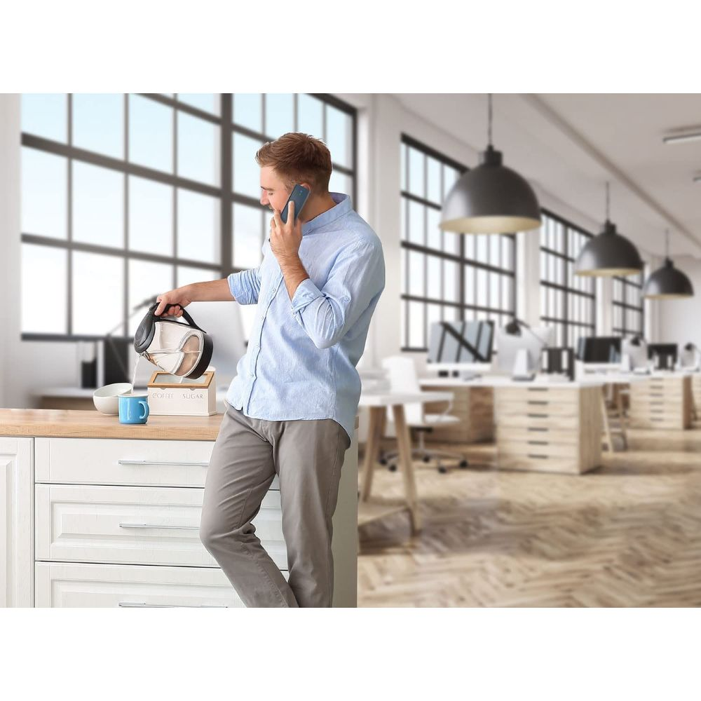 Mann gießt Wasser aus Wasserkocher in Tasse, telefoniert im Büro.