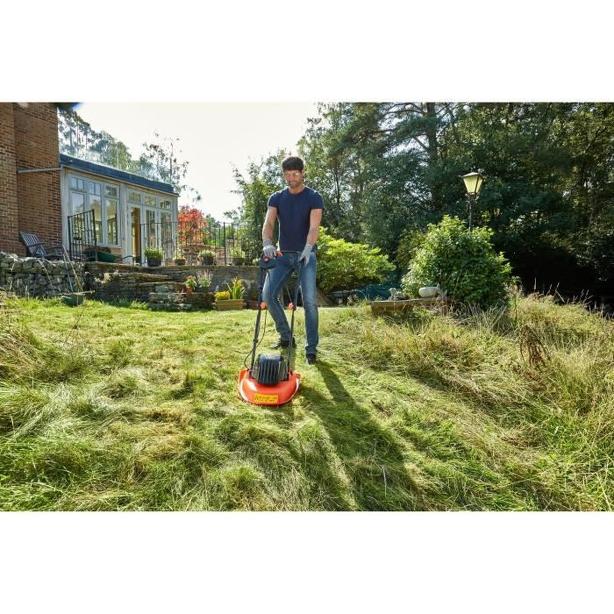 Mann mäht Rasen mit orangenem Rasenmäher vor einem Haus mit Garten.