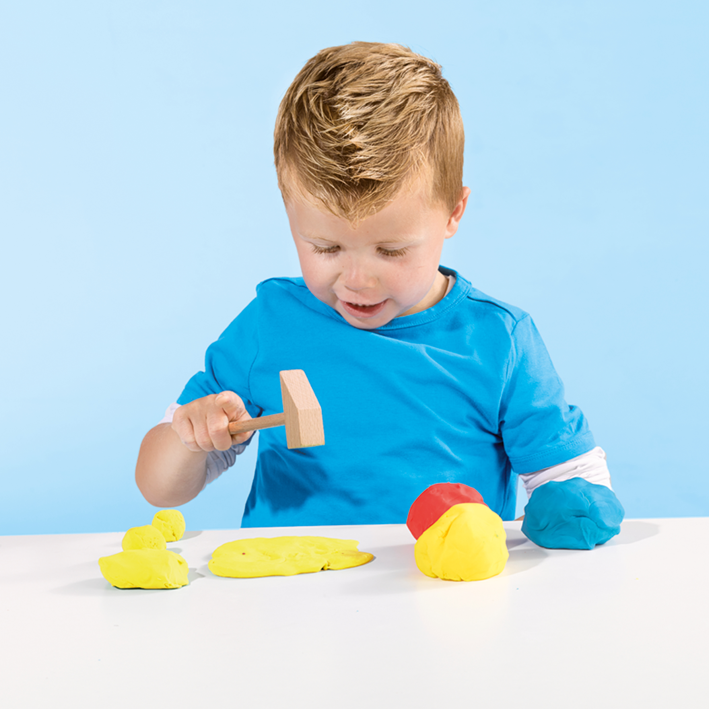 Ein Junge spielt mit Knete und einem Holzhammer vor blauem Hintergrund.