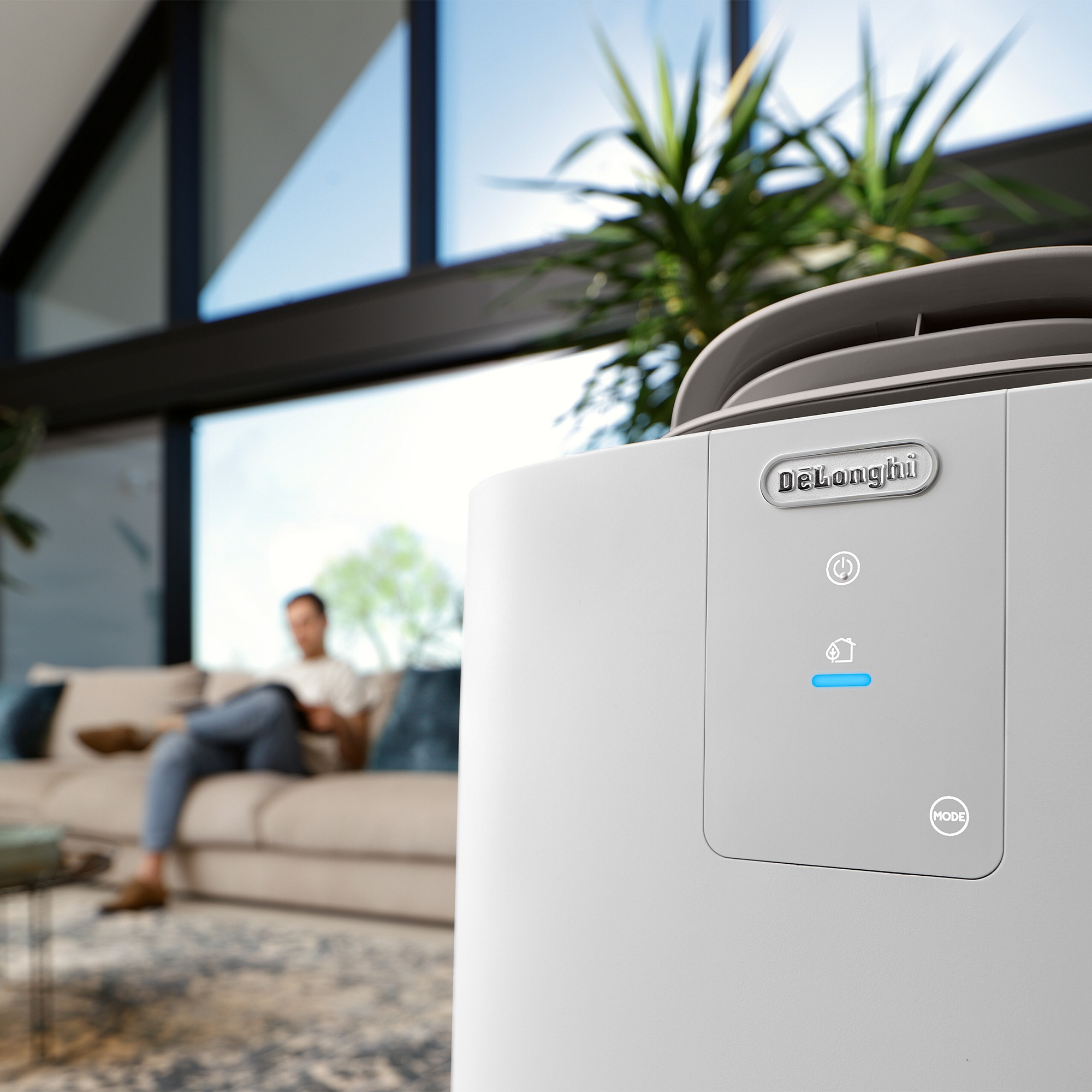 Un purificatore d'aria DeLonghi bianco in un soggiorno moderno. Un uomo legge sul divano.