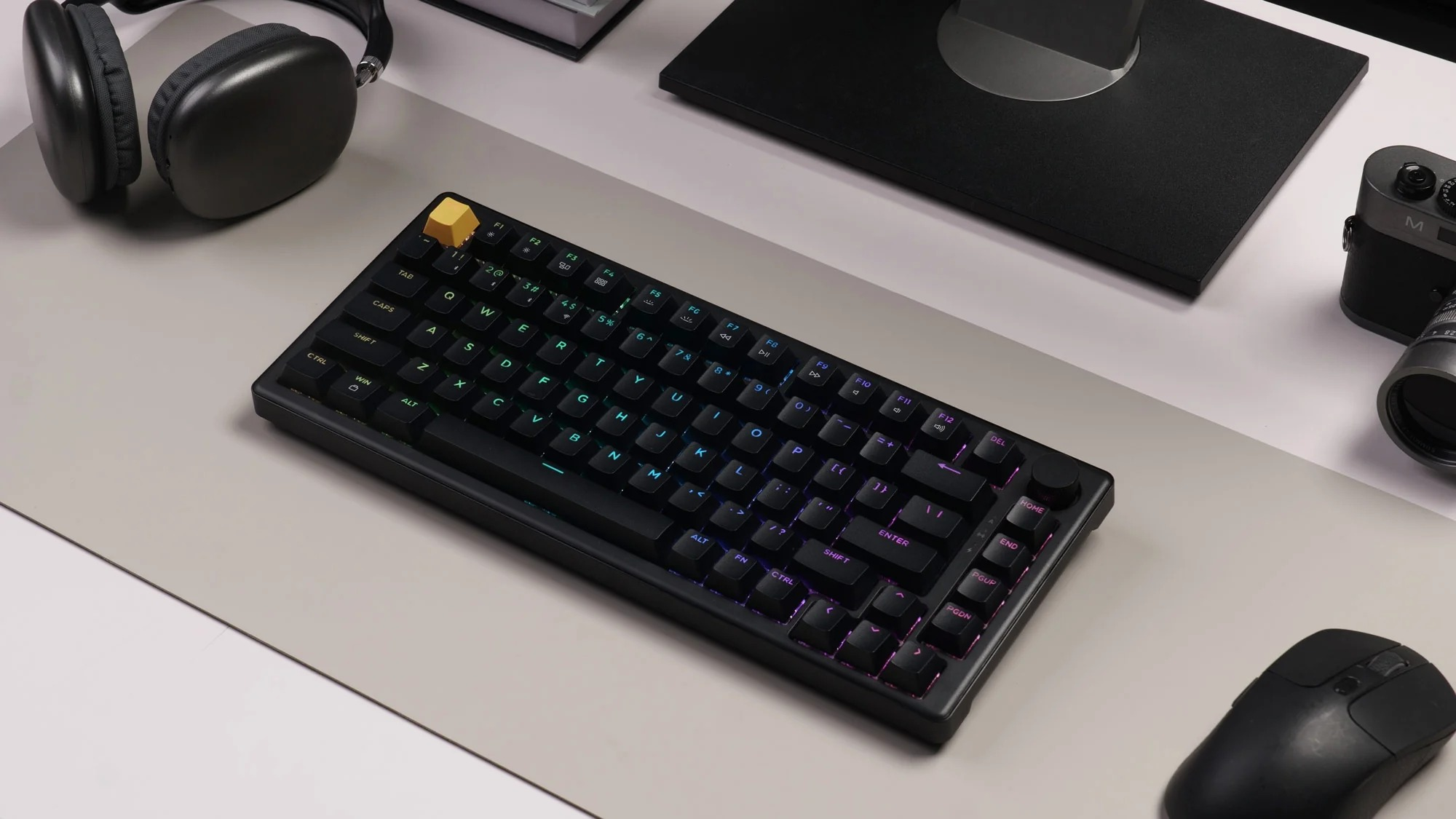 Czarna klawiatura mechaniczna z podświetleniem RGB, słuchawki, mysz i aparat na biurku.