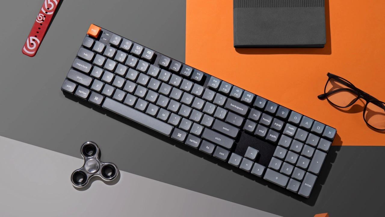 Szara klawiatura z pomarańczowym klawiszem Esc, okularami i fidget spinnerem.