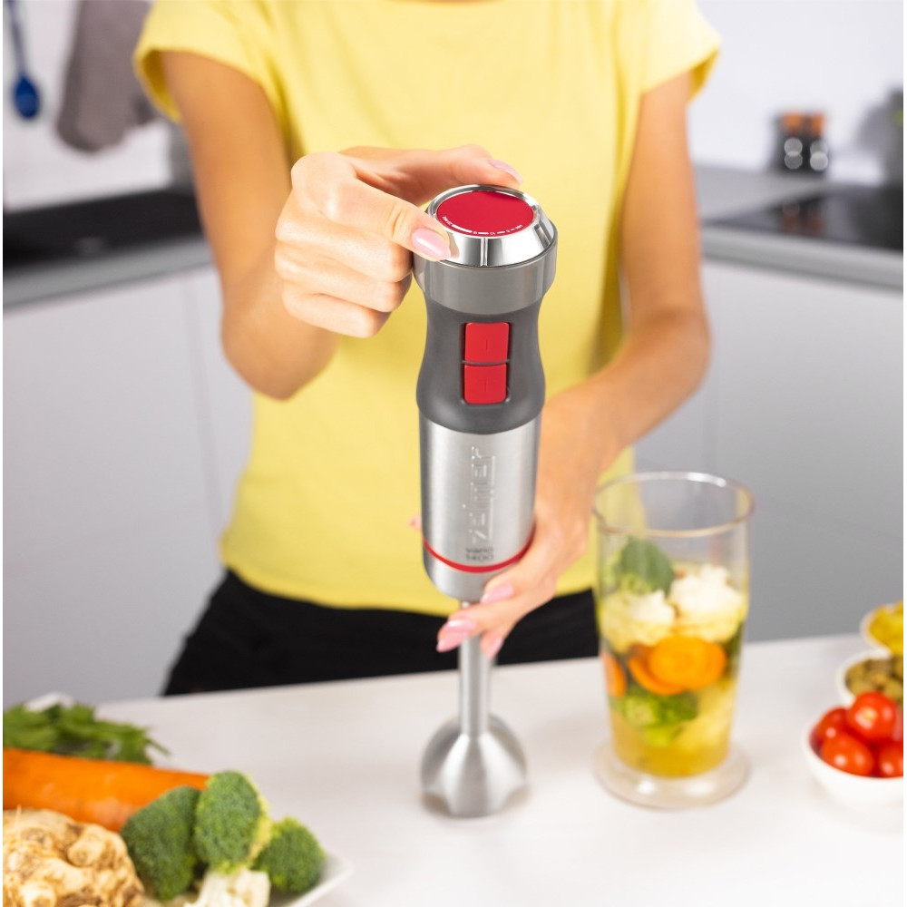 Una mano tiene un frullatore a immersione sopra un bicchiere di verdure in cucina.