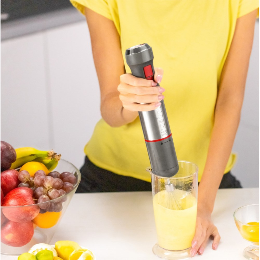 Una persona frulla liquido giallo in un bicchiere con un frullatore a immersione. Frutta vicino.