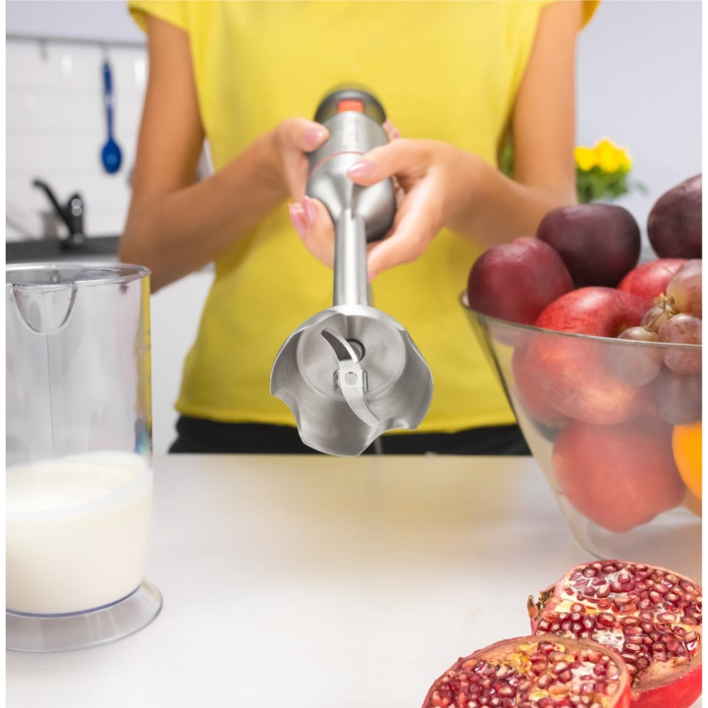 Una persona tiene un frullatore a immersione sopra una tazza di latte, con una ciotola di frutta vicina.
