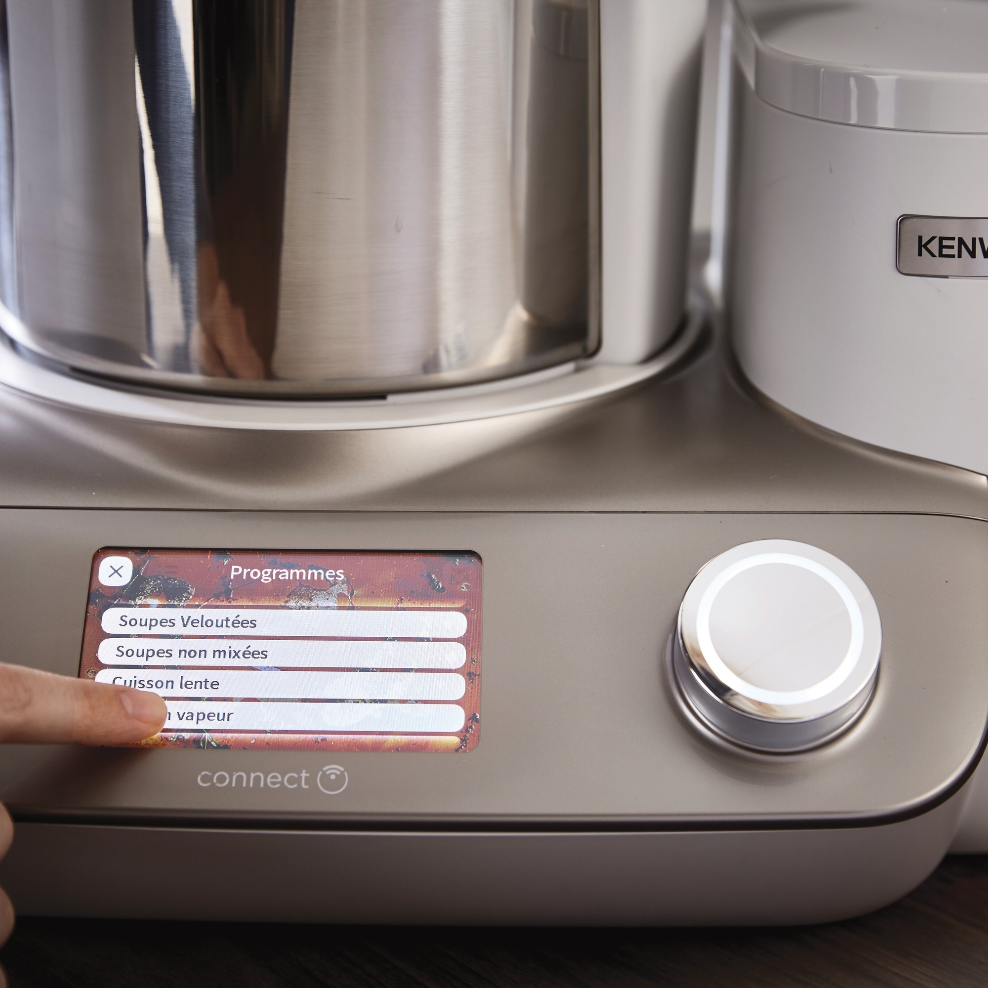 Ein Finger berührt einen Bildschirm mit Kochprogrammen auf einem Kenwood-Gerät.