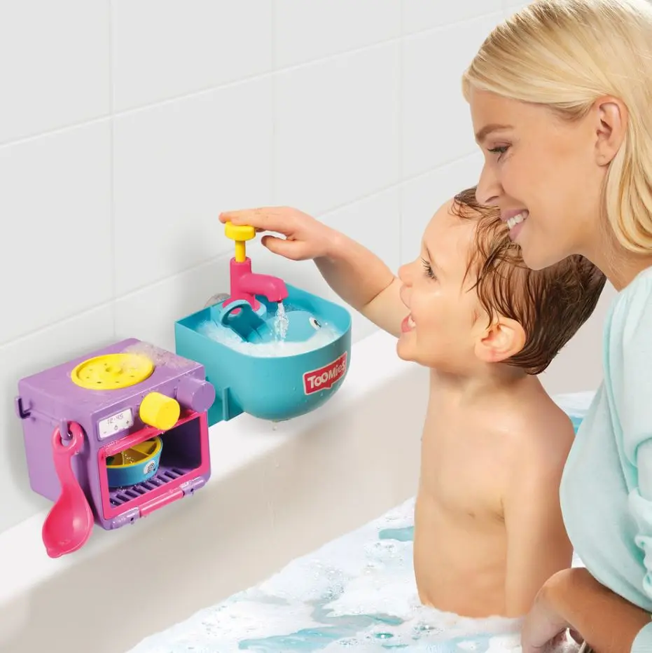 Un niño juega con un lavabo y horno de juguete en una bañera, con su madre observando.