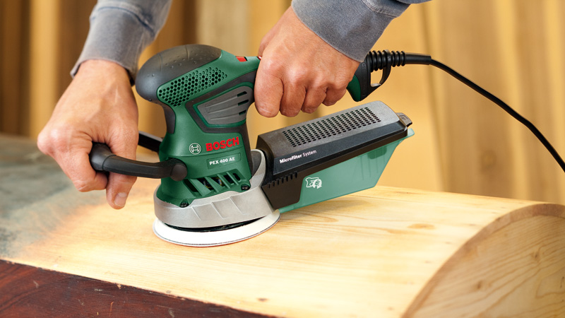 Uomo leviga legno con levigatrice orbitale Bosch verde con raccoglipolvere.