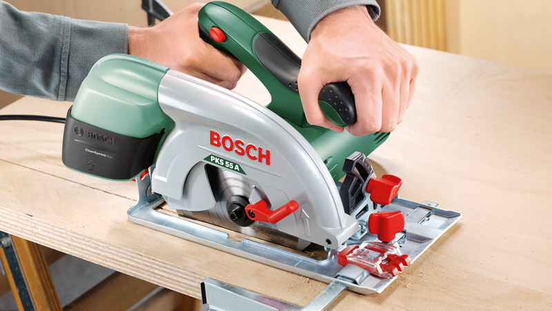 Hände bedienen eine grüne Bosch Kreissäge, die Holz schneidet.
