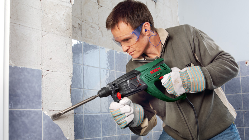 Hombre con gafas y guantes usa taladro percutor para quitar azulejos de pared.