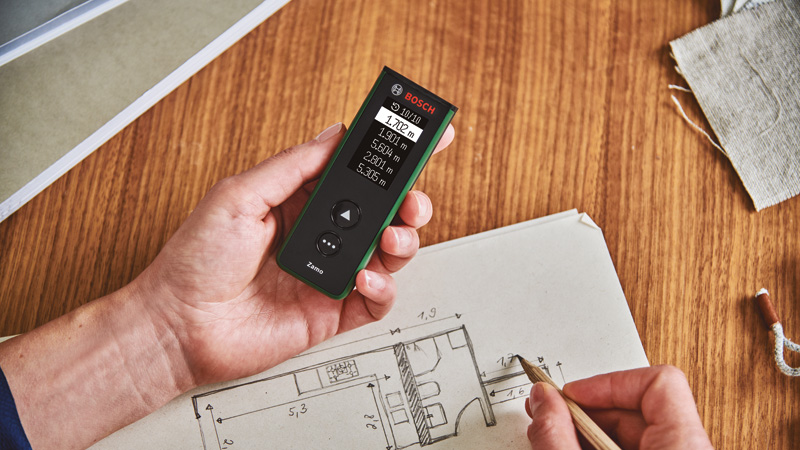 Eine Hand hält ein grünes Bosch-Messgerät über einem Bauplan. Ein Bleistift zeichnet auf dem Plan.