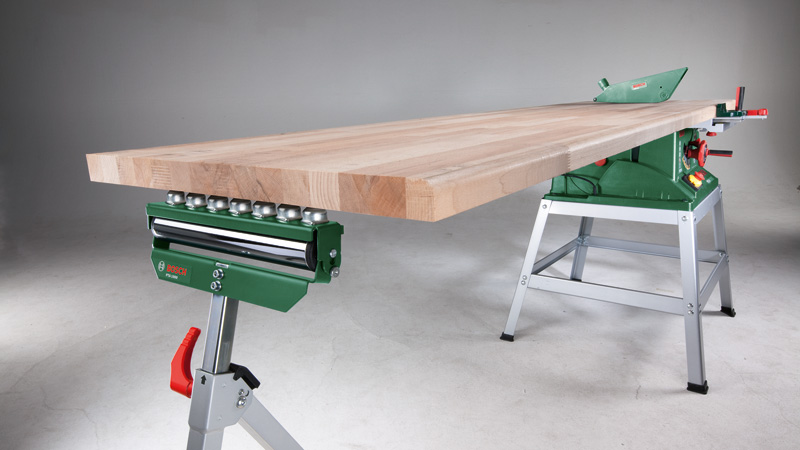 Holzplatte auf grünem Sägetisch mit Rollenhalterung und Untergestell.