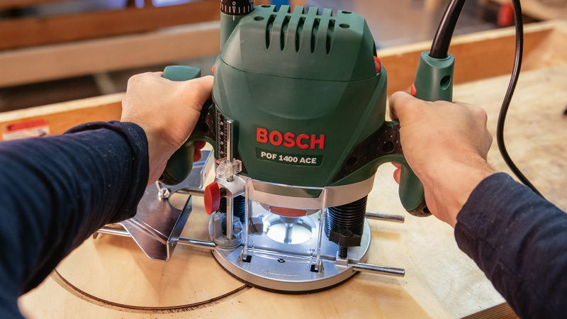 Manos sujetando una fresadora Bosch verde sobre madera.