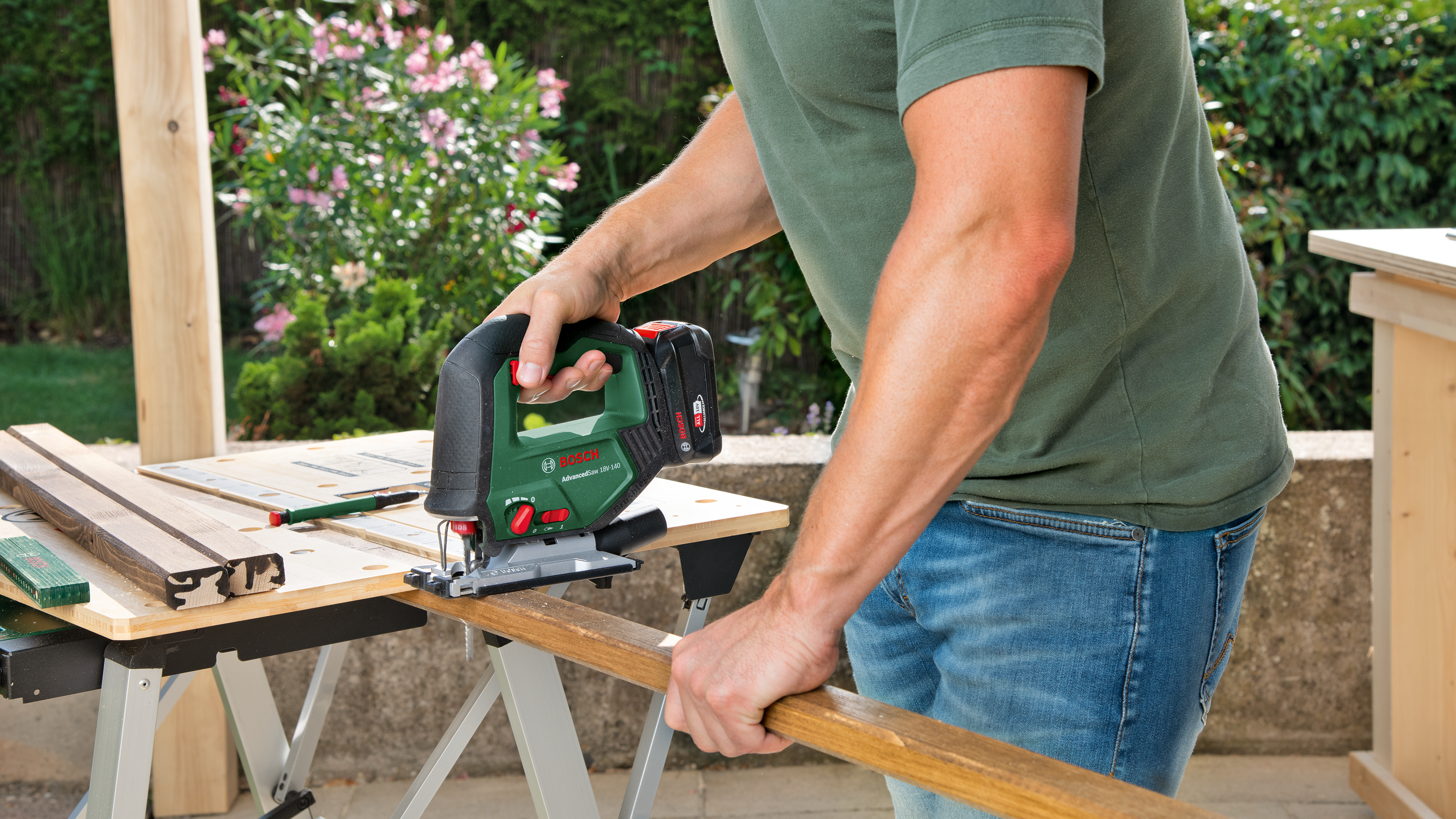 Mann schneidet Holz mit Stichsäge im Garten. Werkbank und Blumen im Hintergrund.