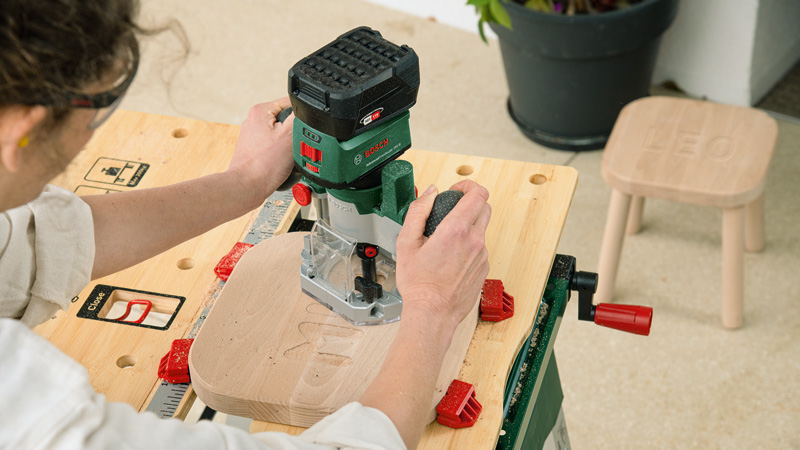Person bearbeitet Holz mit Oberfräse auf Werkbank; kleiner Hocker im Hintergrund.