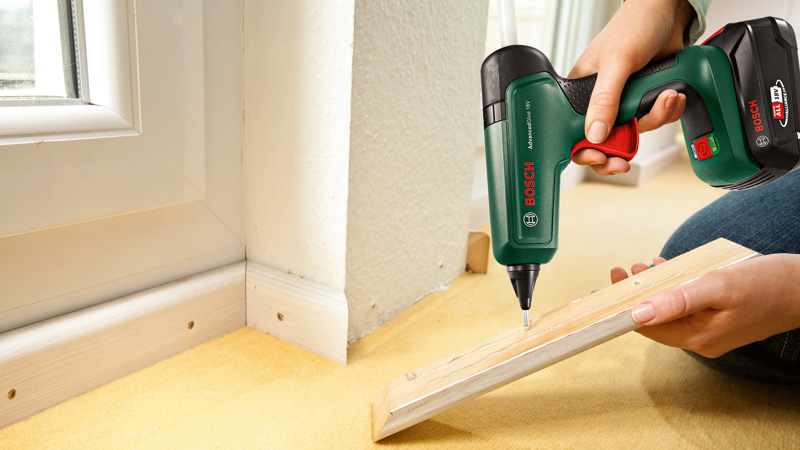 Person benutzt einen grünen Bosch-Klebestift an einem Holzstück neben einer Wand und Fensterecke.