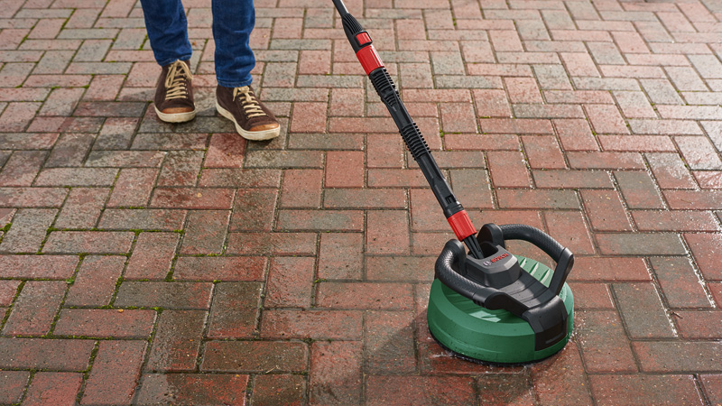 Persona pulisce patio in mattoni rossi con idropulitrice verde.