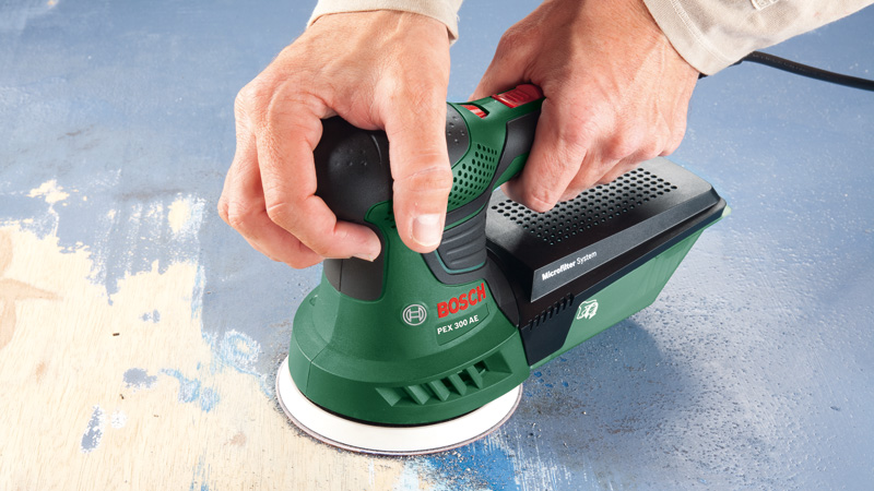 Lijadora orbital Bosch verde con caja de recogida de polvo sobre superficie azul.