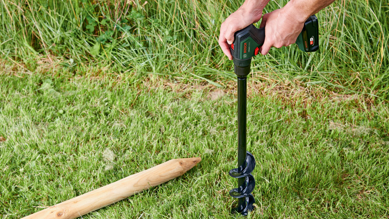 Eine Person benutzt einen Erdbohrer neben einem Holzpfahl im Gras.