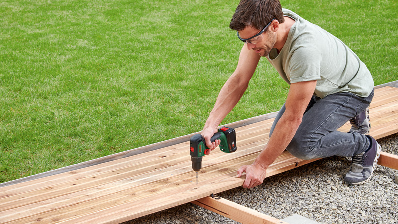 Mann bohrt Holzplanken für eine Terrasse auf einer Schotterfläche.