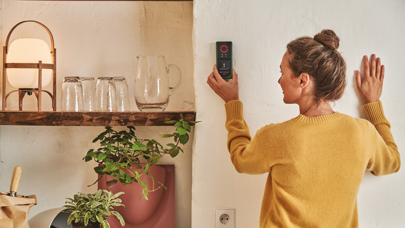 Frau mit gelbem Pullover hält Messgerät an weiße Wand. Regal mit Lampe und Gläsern.