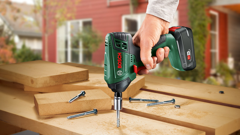 Mano che tiene trapano Bosch verde, avvitando in assi di legno. Viti appoggiate sul tavolo.