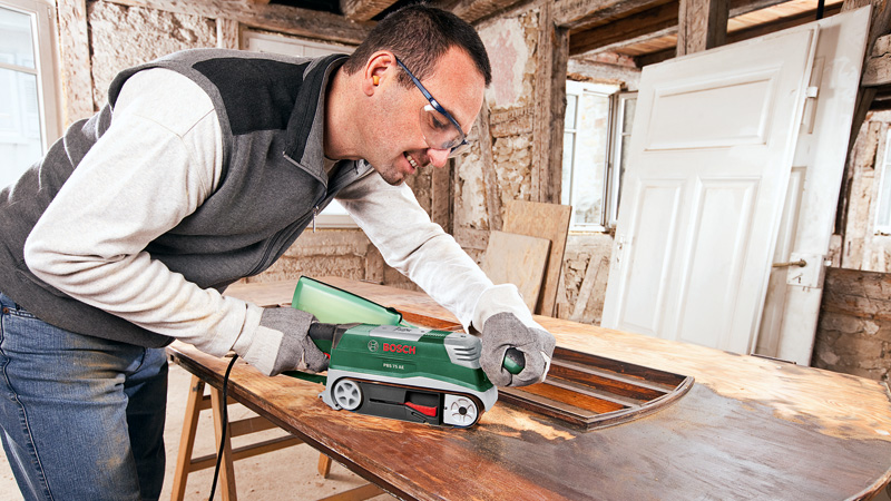 Hombre lijando una mesa de madera con una lijadora de banda Bosch en una habitación rústica.