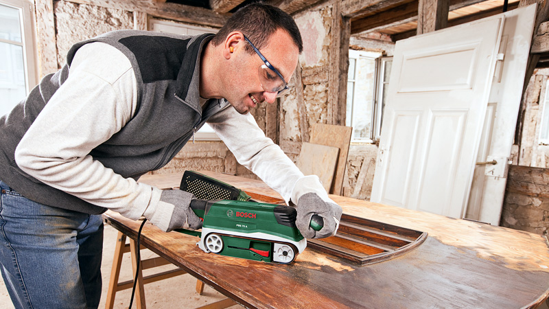 Hombre lijando tablero de mesa con lijadora de banda en taller con puertas.