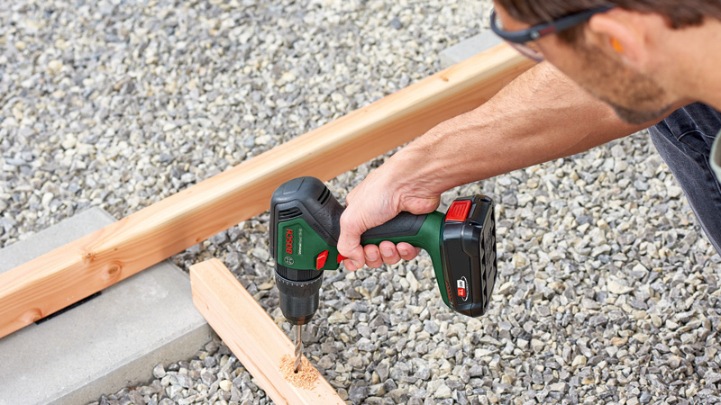 Un hombre taladra madera con un taladro inalámbrico Bosch verde sobre grava.