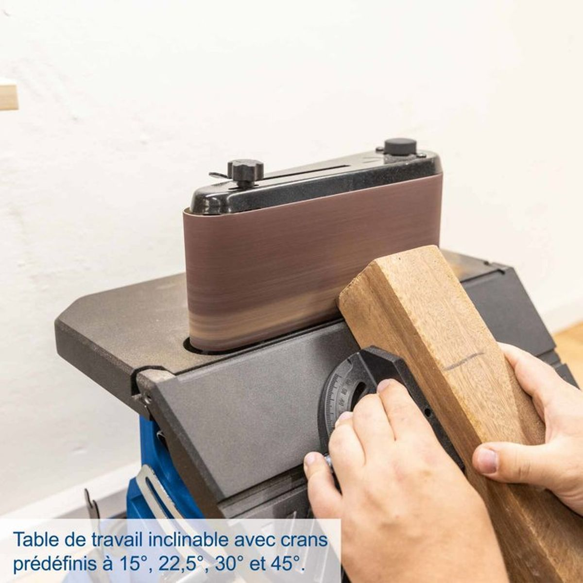 Eine Hand hält ein Holzstück gegen einen Schleifbandschleifer mit verstellbarem Tisch.