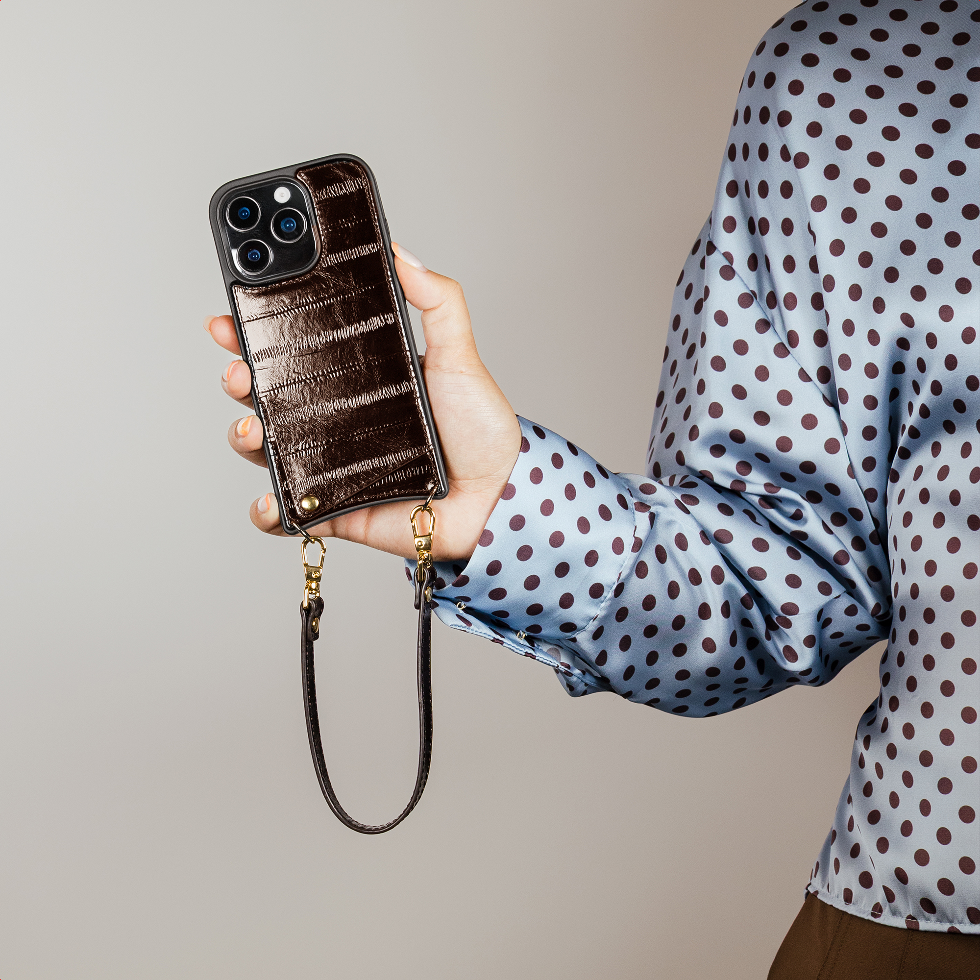 A hand holds a dark brown phone case with a gold strap attached. The person wears a blue polka dot shirt.