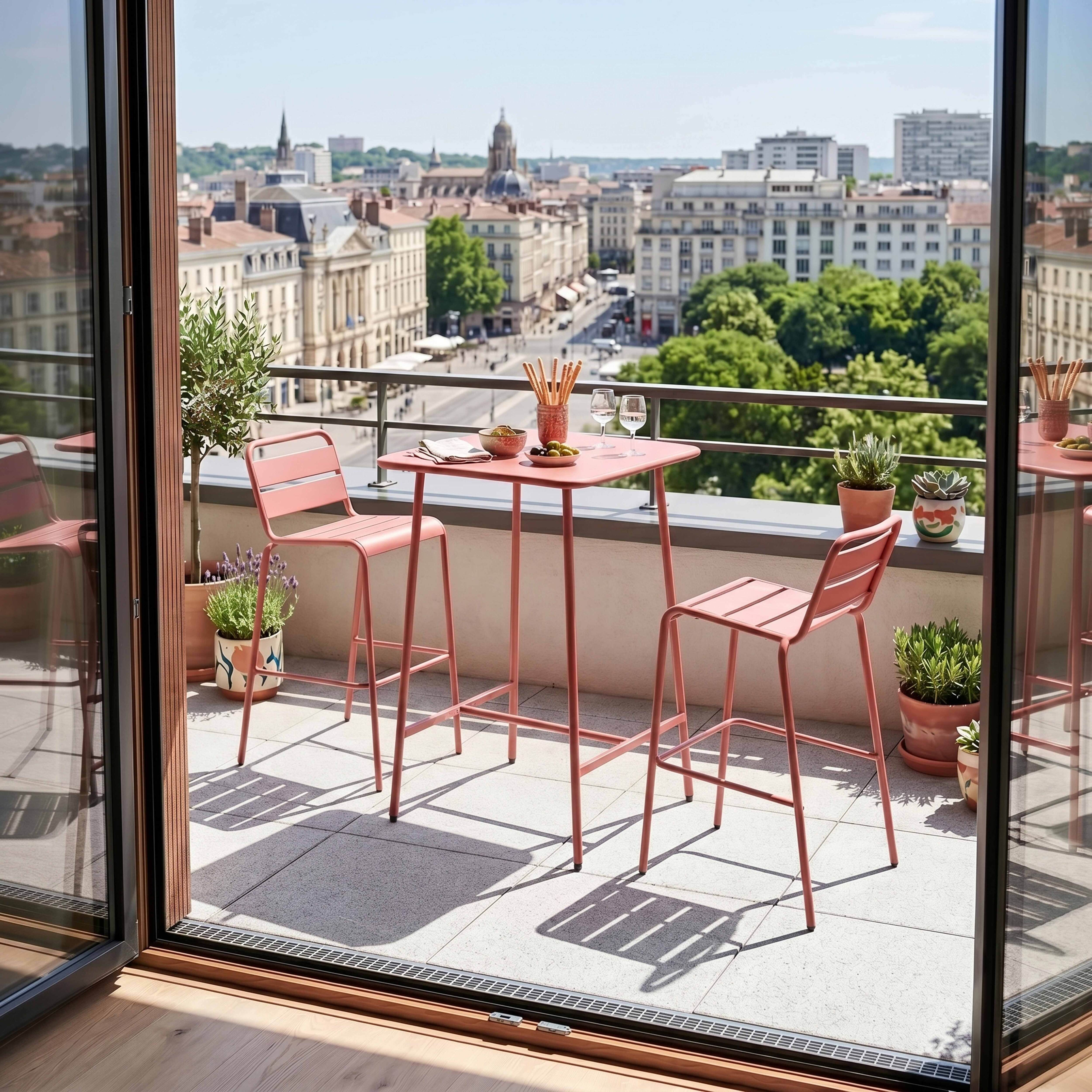 Ein rosafarbener Bistrotisch mit zwei Hockern auf einem Balkon mit Stadtblick.