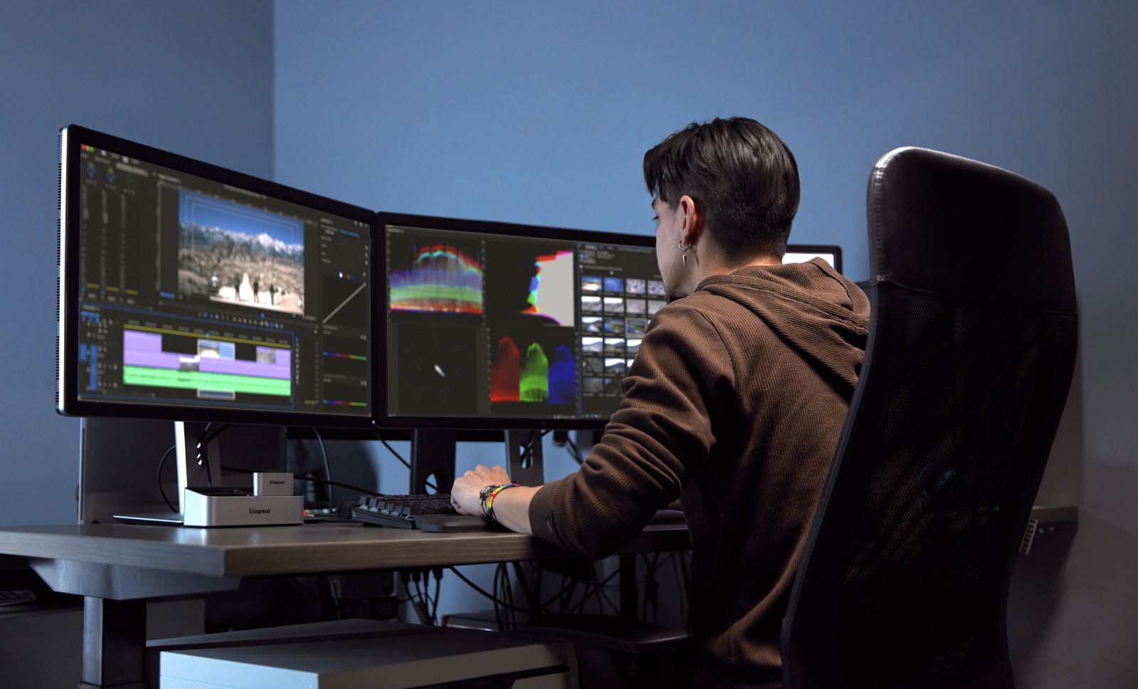 Una persona seduta davanti a tre monitor di computer che mostrano software di editing video.