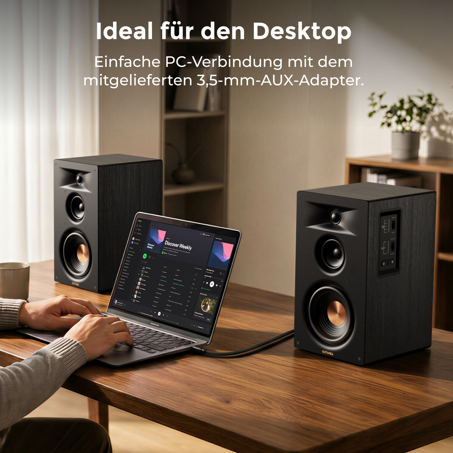 Zwei schwarze Lautsprecher stehen auf einem Holztisch neben einem Laptop. Eine Person tippt auf dem Laptop.
