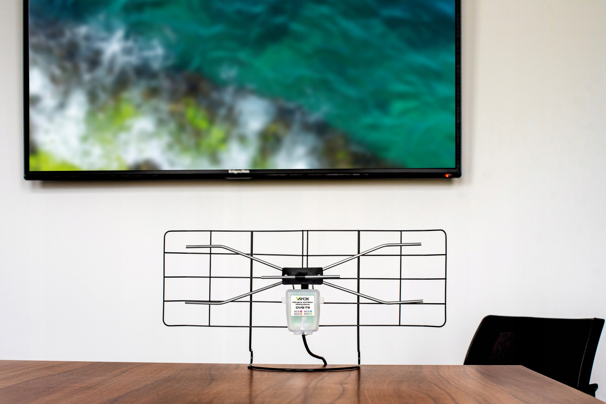 Ein schwarzer Indoor-TV-Antennenständer steht auf einem Holztisch vor einem verschwommenen Fernsehbildschirm.